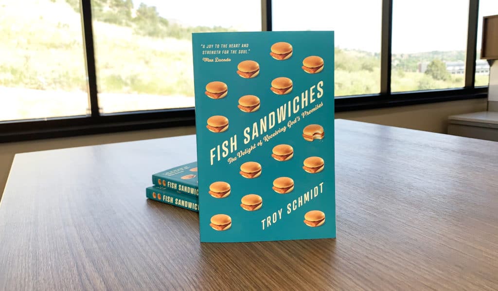 Three copies of "Fish Sandwiches", a book by Troy Schmidt with a blue cover, sitting on a table with a trees in the window behind them.