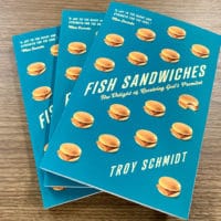 Three copies of "Fish Sandwiches", a book by Troy Schmidt with a blue cover, sitting on a table with a trees in the window behind them.