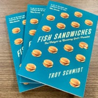 Three copies of "Fish Sandwiches", a book by Troy Schmidt with a blue cover, sitting on a table with a trees in the window behind them.