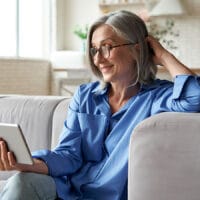 Women’s Bible Study: Living for What Really Matters | Navigators Bible Study Resource | Happy 60s older mature middle aged adult woman holding digital tablet computer conference calling by social distance virtual family online chat meeting or watching video sitting on couch at home.