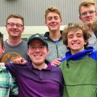 Matthew and his 6 friends are posing for a picture outside of a grey building.