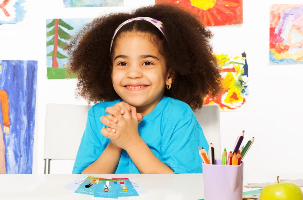 Topical Memory System for Kids | Navigators Bible Study Resource | Happy young girl holds her hands together while sitting in playroom with wall behind full of children drawings