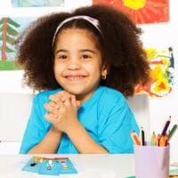 Topical Memory System for Kids | Navigators Bible Study Resource | Happy young girl holds her hands together while sitting in playroom with wall behind full of children drawings