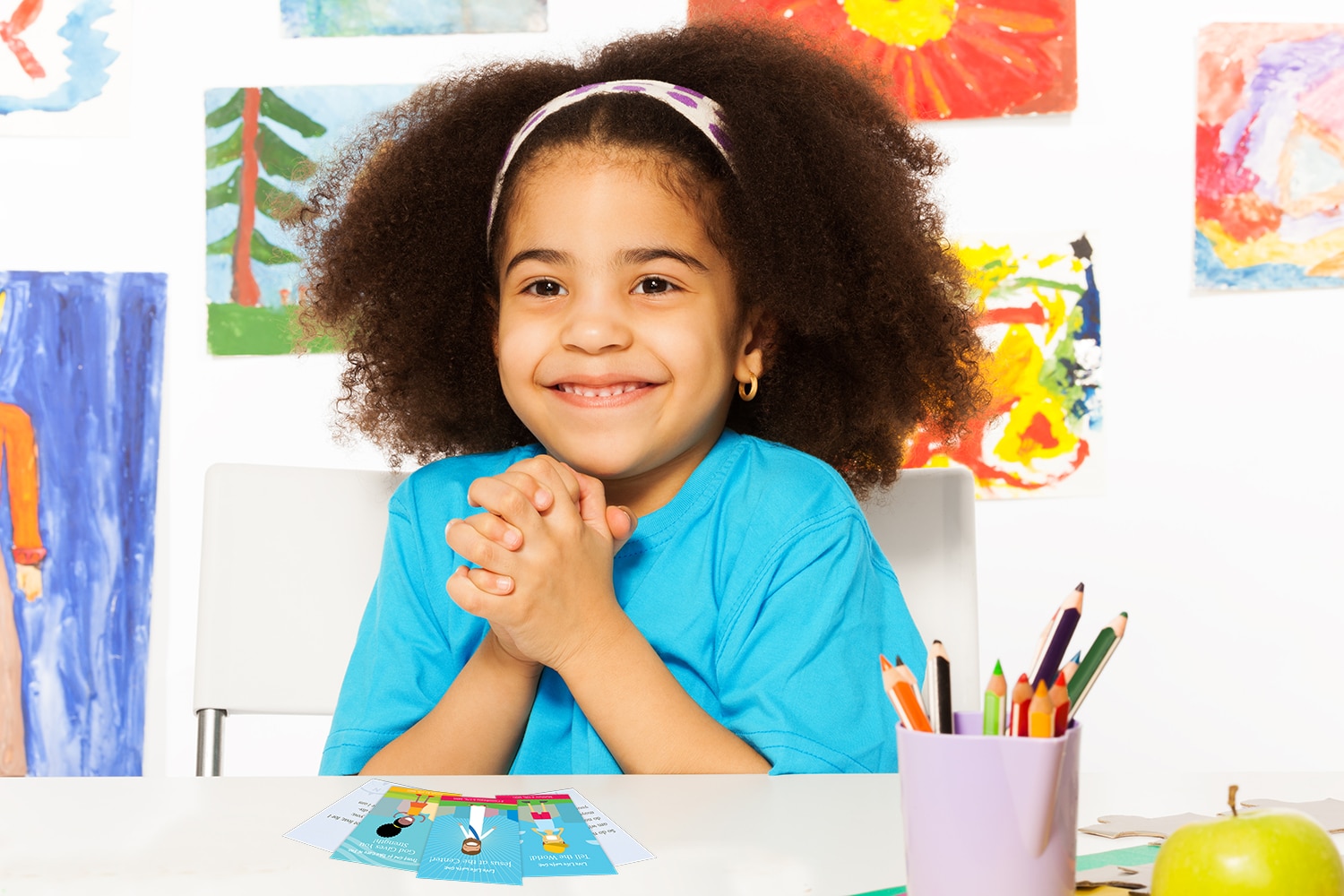 Topical Memory System for Kids | Navigators Bible Study Resource | Happy young girl holds her hands together while sitting in playroom with wall behind full of children drawings