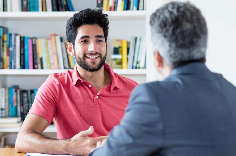 Two Middle-Eastern men talking at a table in a library, with a shelf of books behind them.