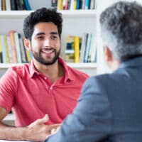 Two Middle-Eastern men talking at a table in a library, with a shelf of books behind them.