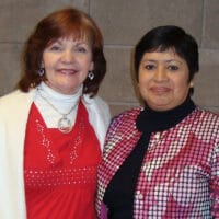 Diane and Juanita, two Hispanic women, pose for a camera in front of a brick wall.