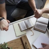 Parents holding hands with their teenage son praying together with bibles open on the table in front of them.