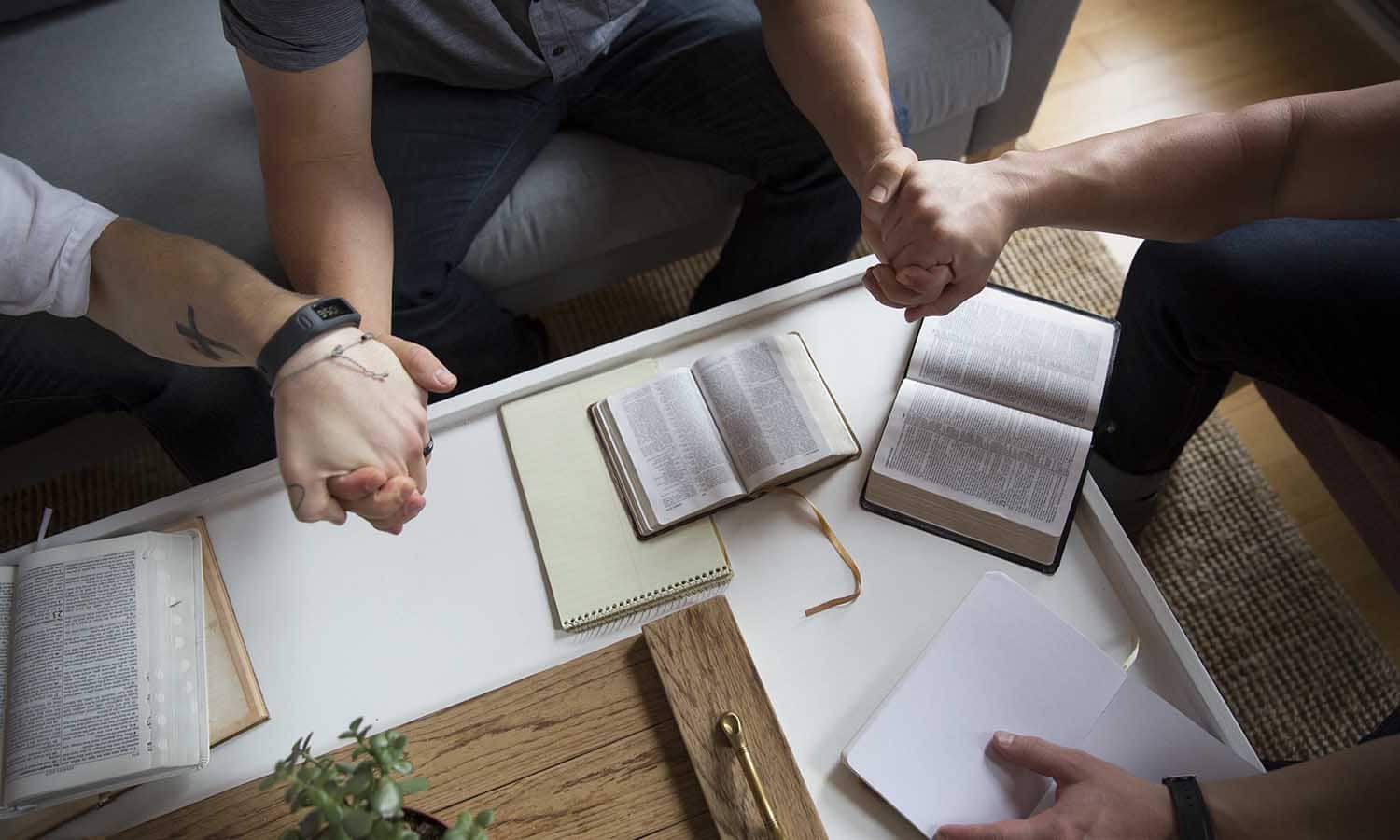 Parents holding hands with their teenage son praying together with bibles open on the table in front of them.