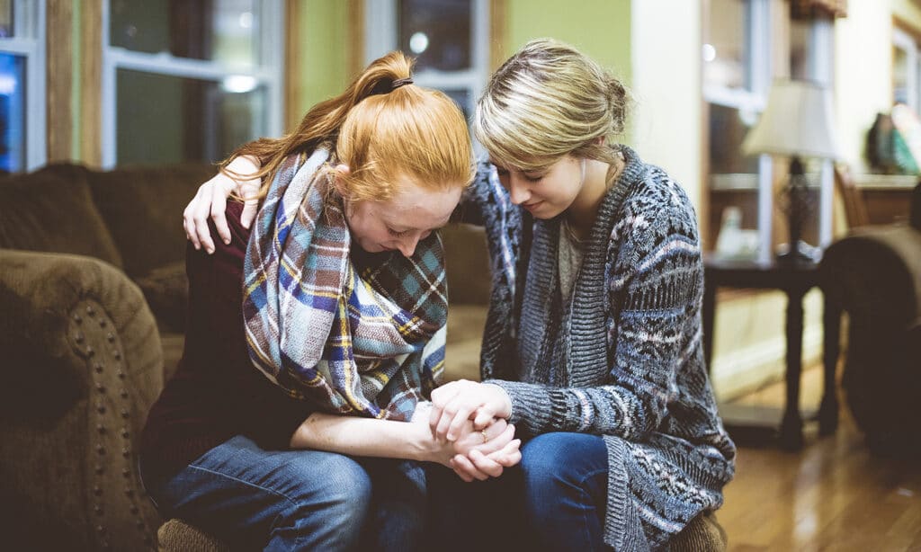 Two women in a sorority praying together on a couch inside of a house.