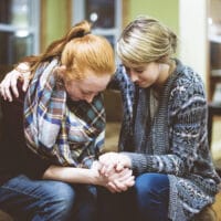 Two women in a sorority praying together on a couch inside of a house.