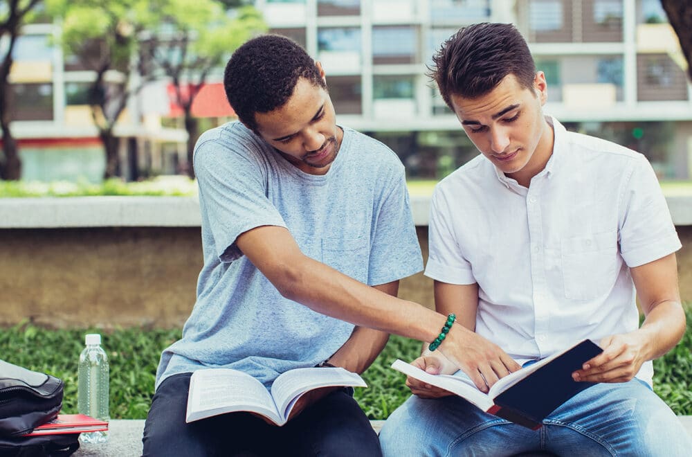 Two young men read their bibles together while sitting out on a lawn in front of a dorm building.