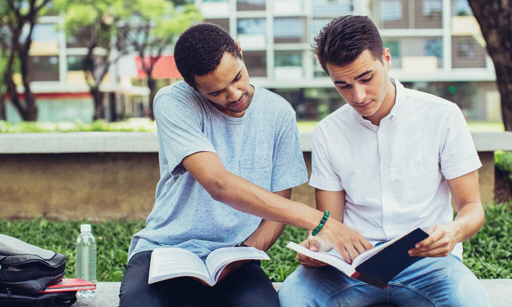 Two young men read their bibles together while sitting out on a lawn in front of a dorm building.