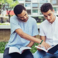 Two young men read their bibles together while sitting out on a lawn in front of a dorm building.