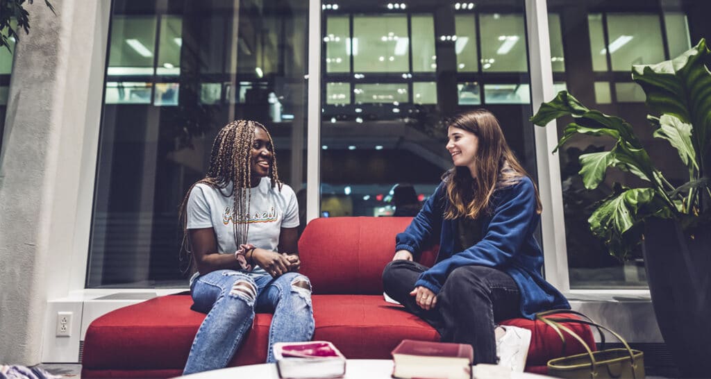 Dierdre and Mia sit on a red couch in front of a large window at night in a common area talking about the benefits of discipleship.