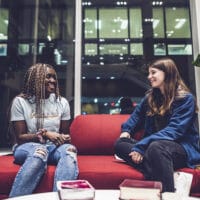 Dierdre and Mia sit on a red couch in front of a large window at night in a common area talking about the benefits of discipleship.