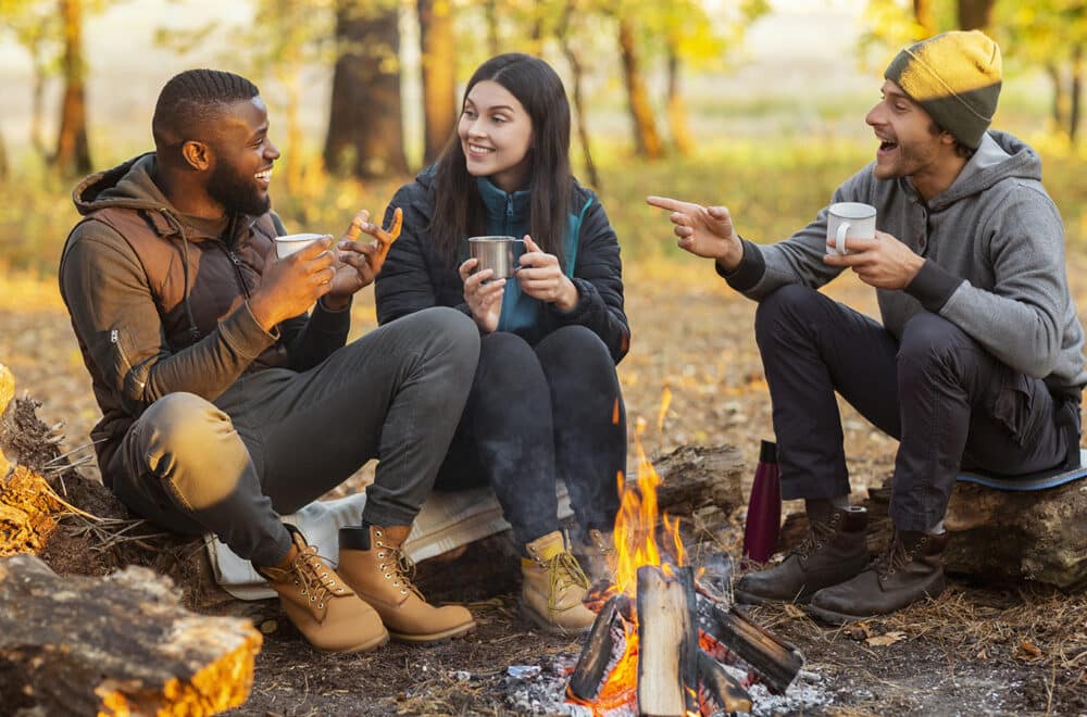 Three young adults sitting around a campfire drinking coffee and practicing thankful prayers.