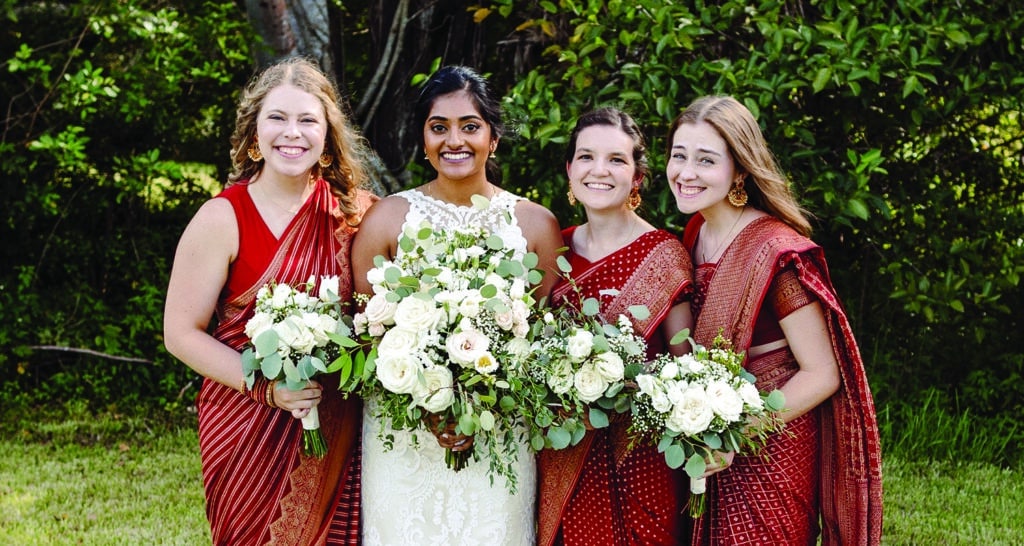 Four women, in dresses, holding flowers and posing in front trees. The second woman is wearing a wedding dress.