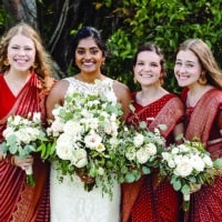 Four women, in dresses, holding flowers and posing in front trees. The second woman is wearing a wedding dress.