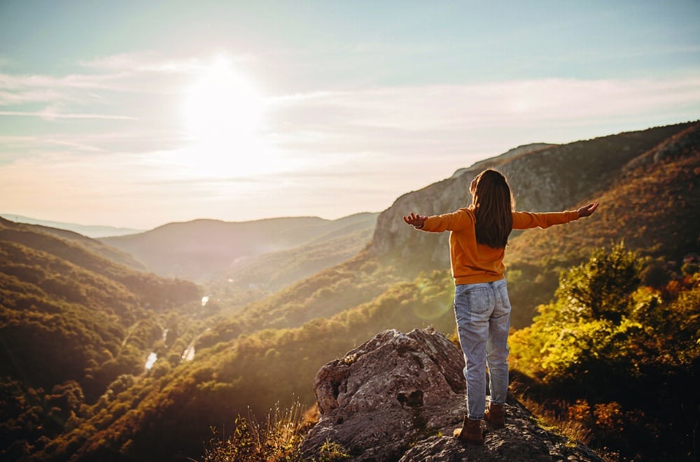 A woman stands on a mountain at sunrise, arms raised in praise and worship. The image embodies the concept of "fruit of the spirit" and the transformative power of faith.