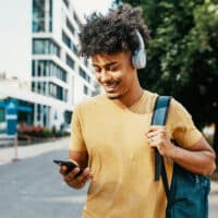 Young Mixed race millennial man listening to a podcast while wearing headphones, walking down a city street.