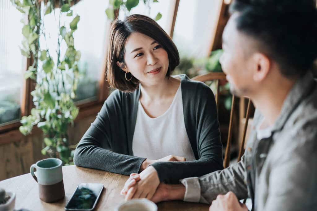 Happy young Asian couple having a coffee date in cafe, holding hands on coffee table. Drinking coffee and chatting. Enjoying a relaxing moment together.