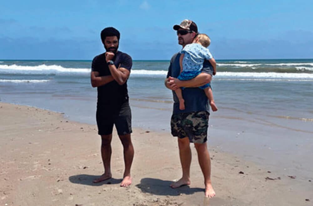 Two men at the beach, chatting by the ocean on the sand. One of the men is holding a baby boy.