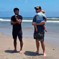 Two men at the beach, chatting by the ocean on the sand. One of the men is holding a baby boy.