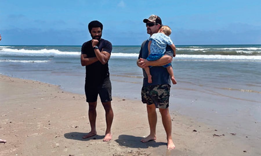 Two men at the beach, chatting by the ocean on the sand. One of the men is holding a baby boy.