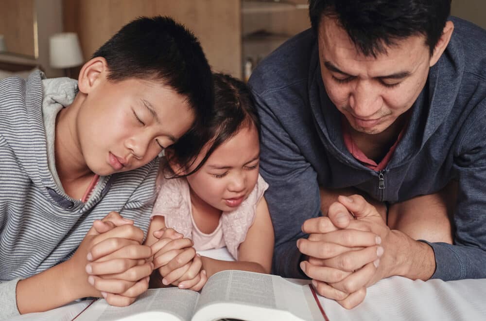 Children and father kneeling in prayer beside a bed with the Bible open in front of them.