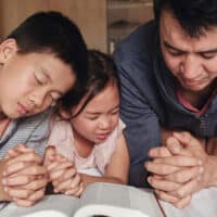 Children and father kneeling in prayer beside a bed with the Bible open in front of them.