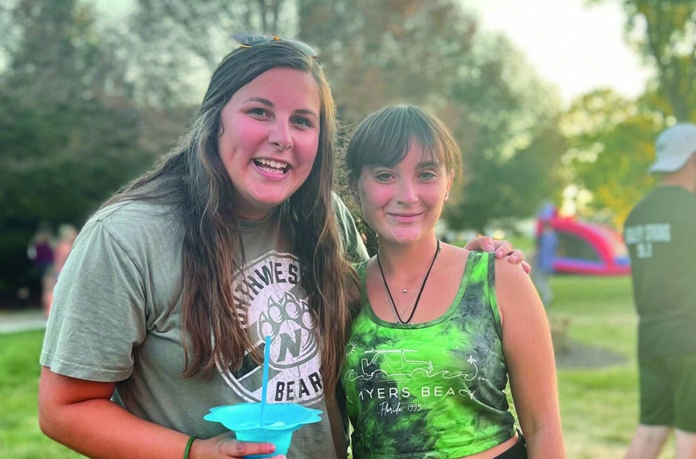 Two young women smiling for the camera as connect at a local festival in a park.