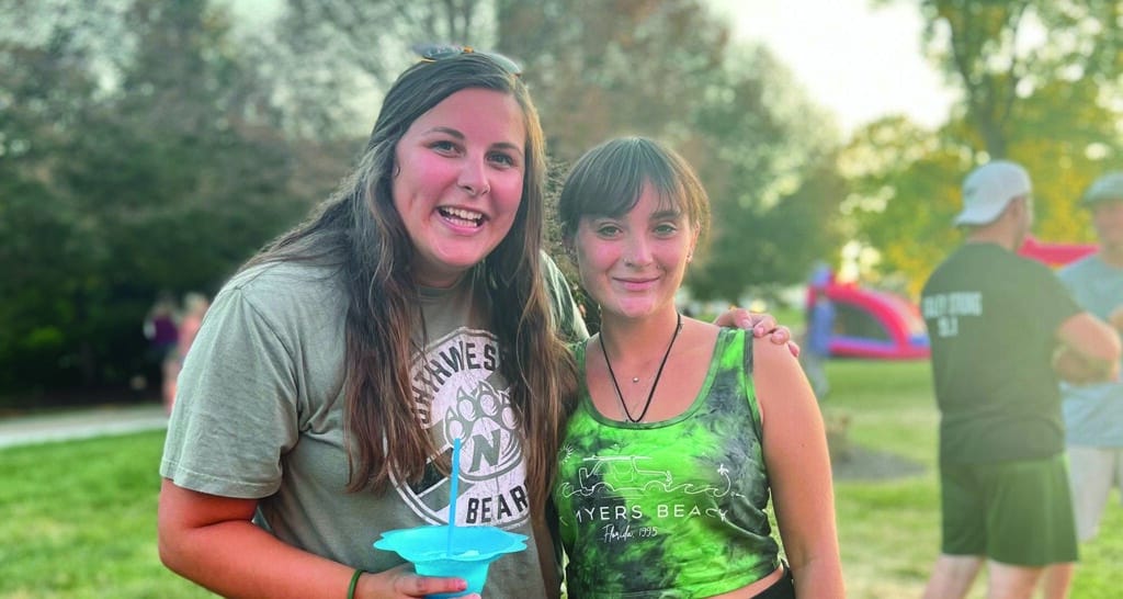 Two young women smiling for the camera as connect at a local festival in a park.