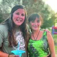 Two young women smiling for the camera as connect at a local festival in a park.