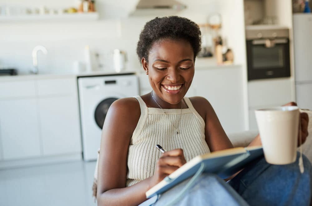 Young woman having tea and writing in a notebook on the sofa at home.