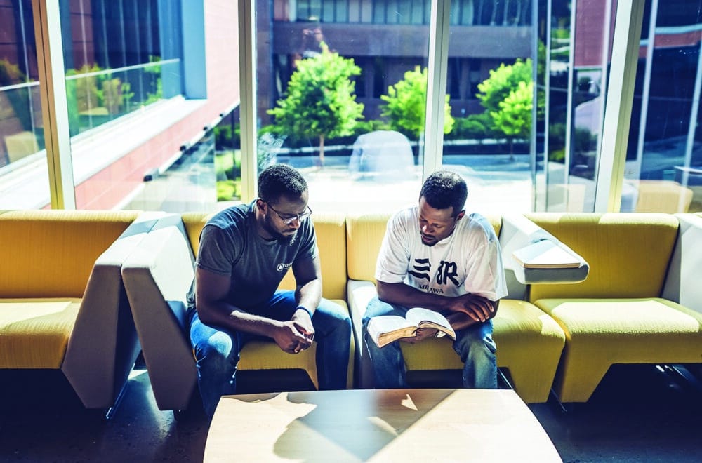 Two African-American men reading the bible together in an office lobby by a window.