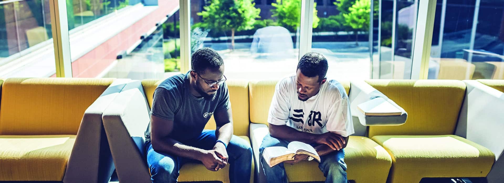 Two African-American men reading the bible together in an office lobby by a window.
