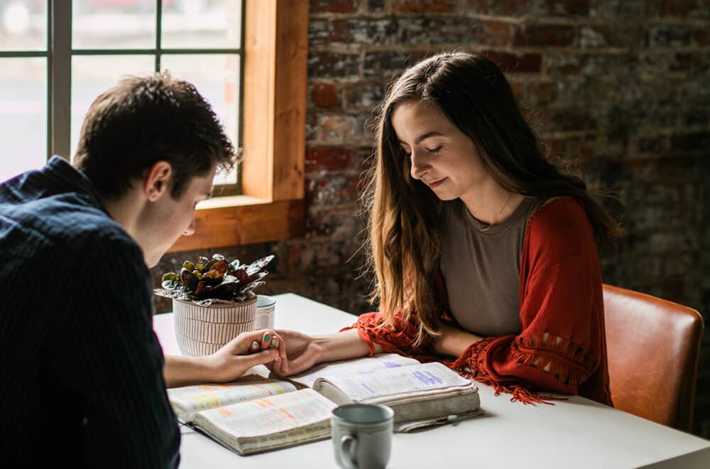 A man and woman sitting across the table from each other praying together and holding hands with their Bibles open on the table.