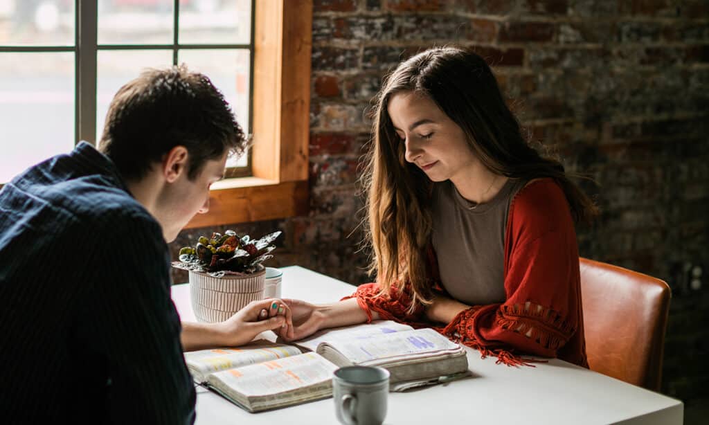 A man and woman sitting across the table from each other praying together and holding hands with their Bibles open on the table.