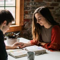 A man and woman sitting across the table from each other praying together and holding hands with their Bibles open on the table.