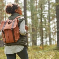 Back view portrait of young African-American woman with backpack enjoying hiking in forest lit by sunlight.