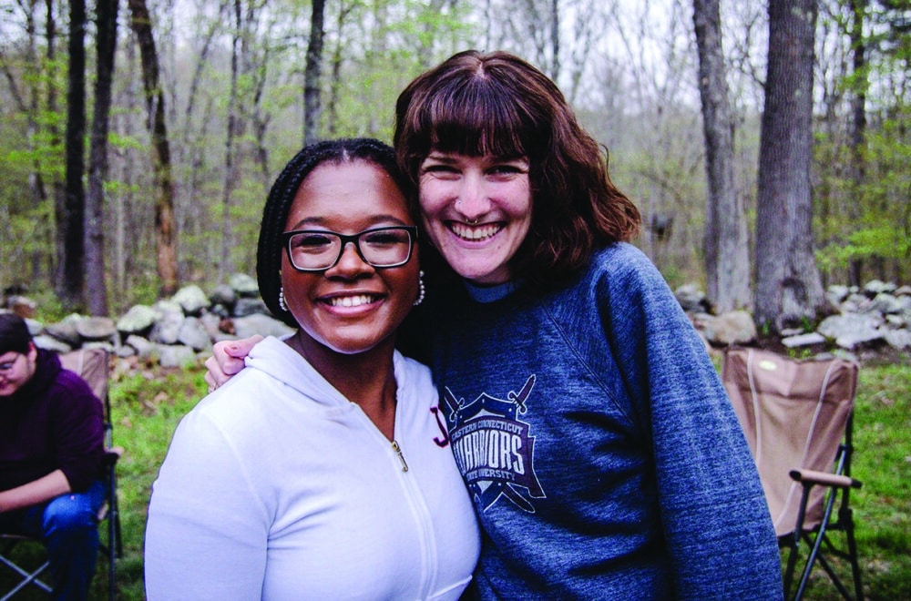 Two college-aged women smiling at a camera in a campground with trees and rocks.