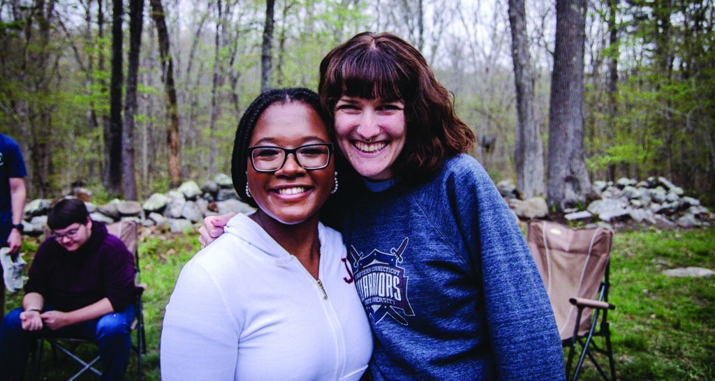 Two college-aged women smiling at a camera in a campground with trees and rocks.