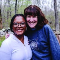 Two college-aged women smiling at a camera in a campground with trees and rocks.