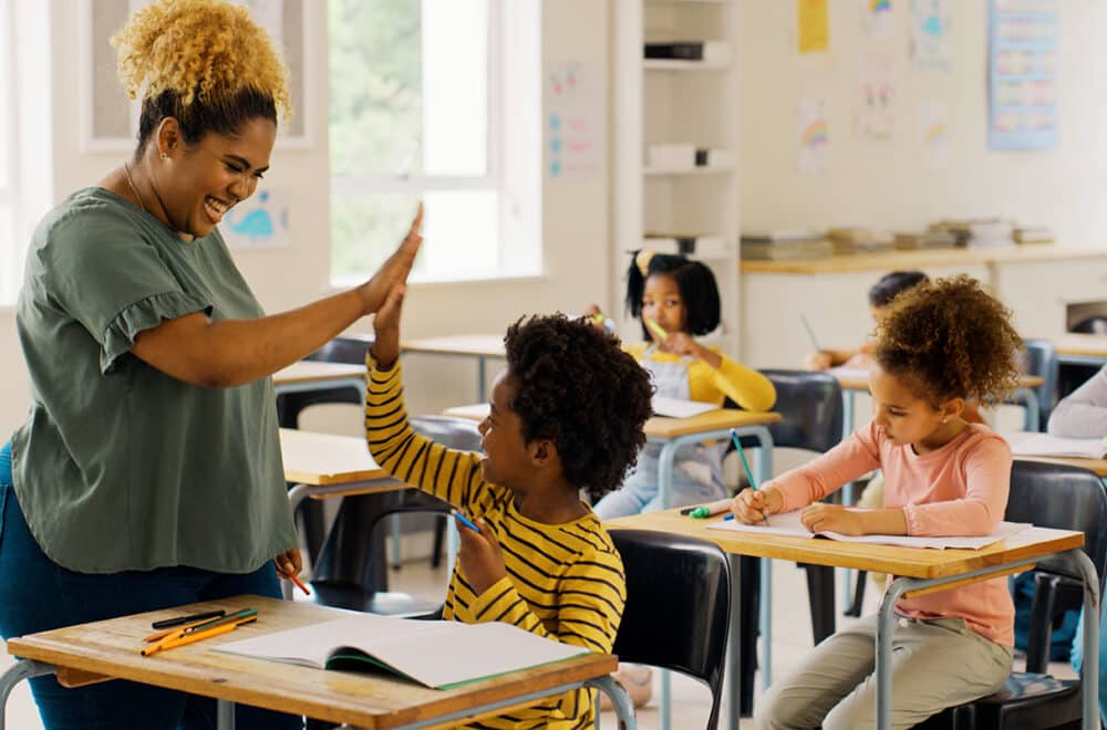 A teacher in a green shirt is giving a high-five to a young student in her class who is sitting at a desk.