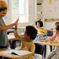 A teacher in a green shirt is giving a high-five to a young student in her class who is sitting at a desk.