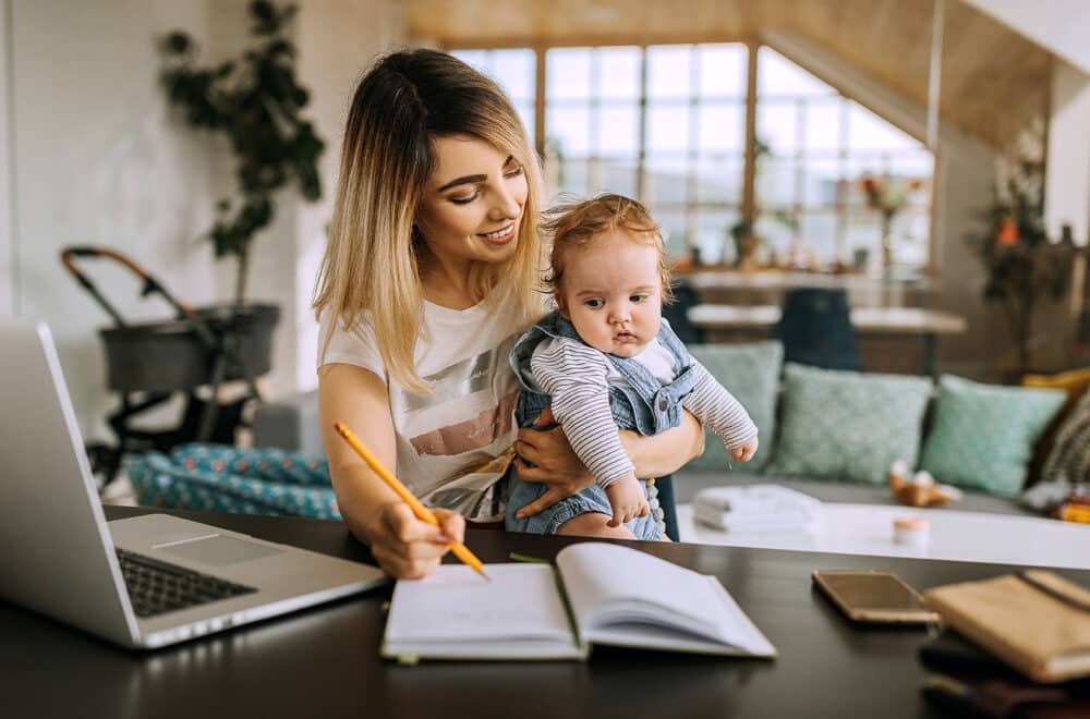 A mother sitting at an office desk in her home while holding her baby and journaling prayers for her child.