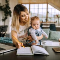 A mother sitting at an office desk in her home while holding her baby and journaling prayers for her child.