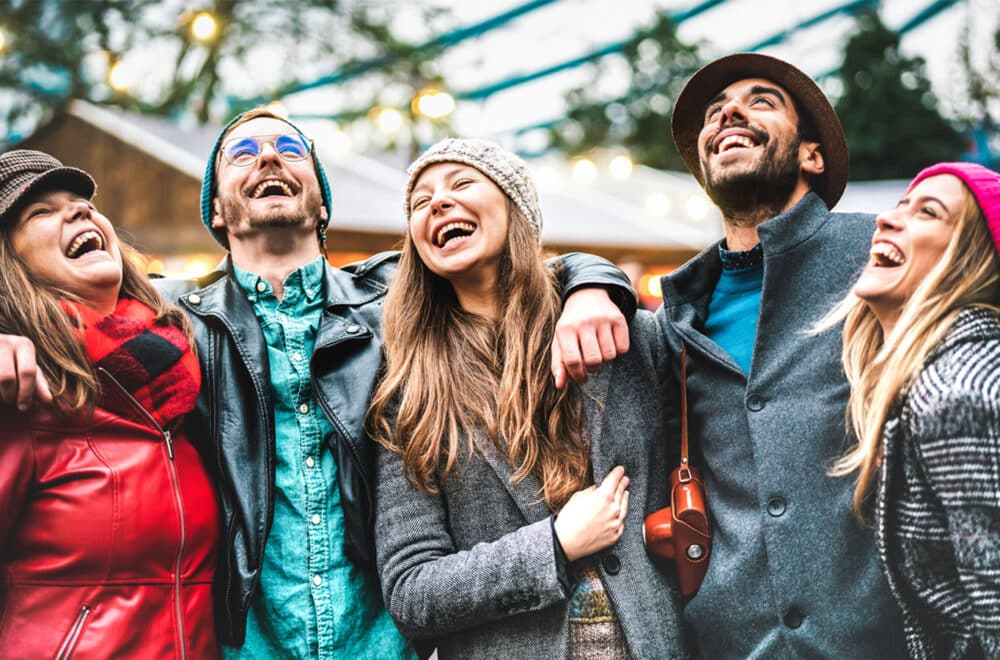 A group of six friends laugh together wearing winter coats outside standing in front of a cabin in the woods.