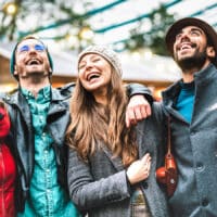 A group of six friends laugh together wearing winter coats outside standing in front of a cabin in the woods.